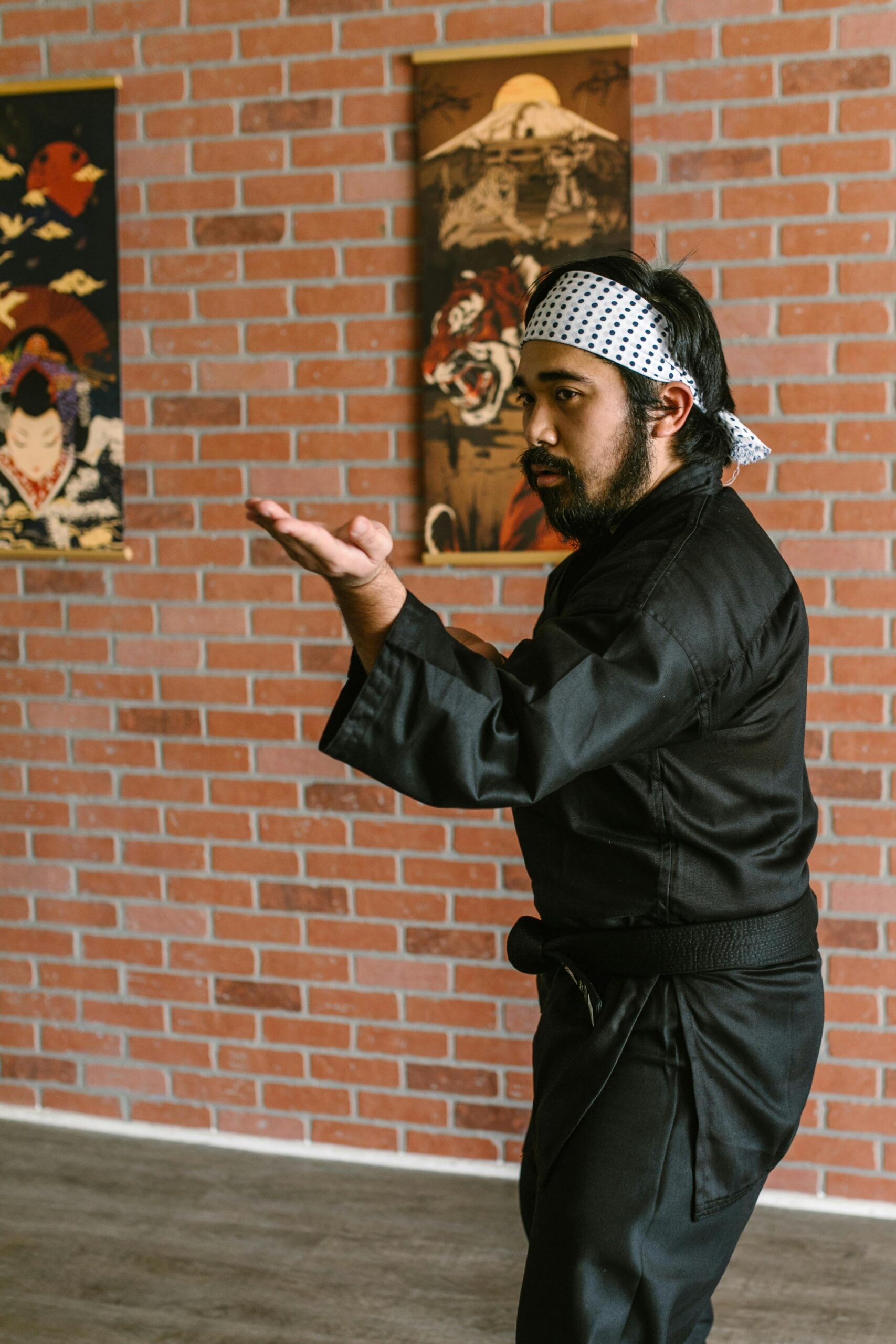 A martial artist in a black gi poses in a dojo against a brick wall backdrop.